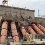 Vista de uma grande usina hidrelétrica, com estruturas industriais, tubulações e linhas de transmissão de energia.