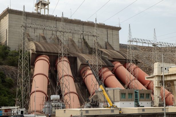 Vista de uma grande usina hidrelétrica, com estruturas industriais, tubulações e linhas de transmissão de energia.