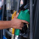 Mão segura um bico verde de abastecimento em um posto de combustíveis.