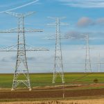 Torres de transmissão de energia elétrica se estendem por campos verdes sob céu azul.