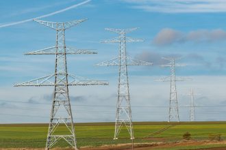 Torres de transmissão de energia elétrica se estendem por campos verdes sob céu azul.