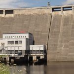 Barragem hidrelétrica junto a um edifício, às margens de um reservatório de águas calmas.