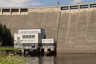 Barragem hidrelétrica junto a um edifício, às margens de um reservatório de águas calmas.
