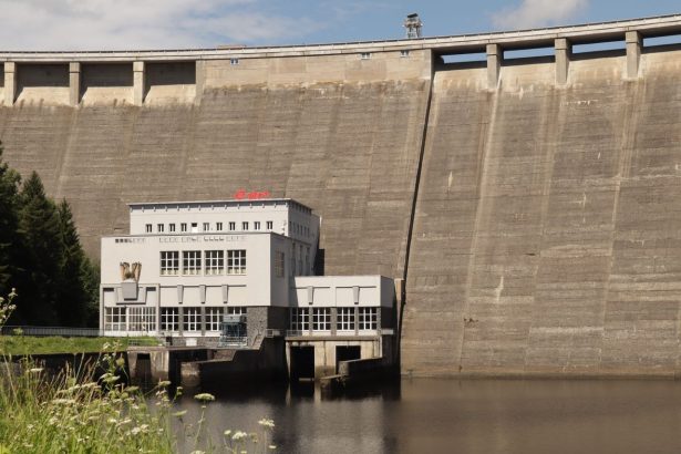 Barragem hidrelétrica junto a um edifício, às margens de um reservatório de águas calmas.