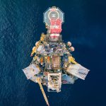 Vista aérea de uma plataforma de petróleo offshore, com heliponto, cercada pelo oceano azul.