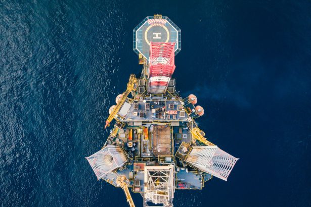 Vista aérea de uma plataforma de petróleo offshore, com heliponto, cercada pelo oceano azul.