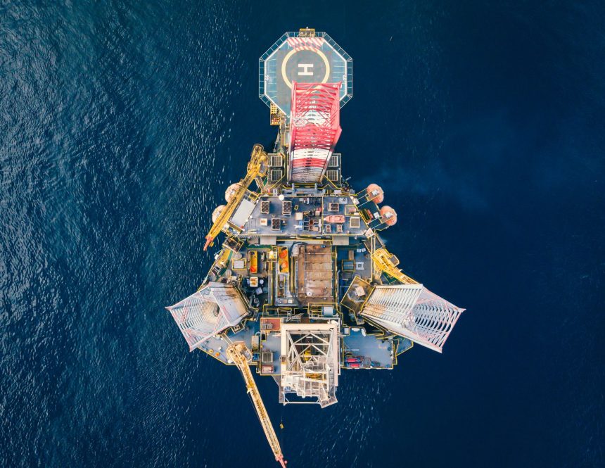 Vista aérea de uma plataforma de petróleo offshore, com heliponto, cercada pelo oceano azul.